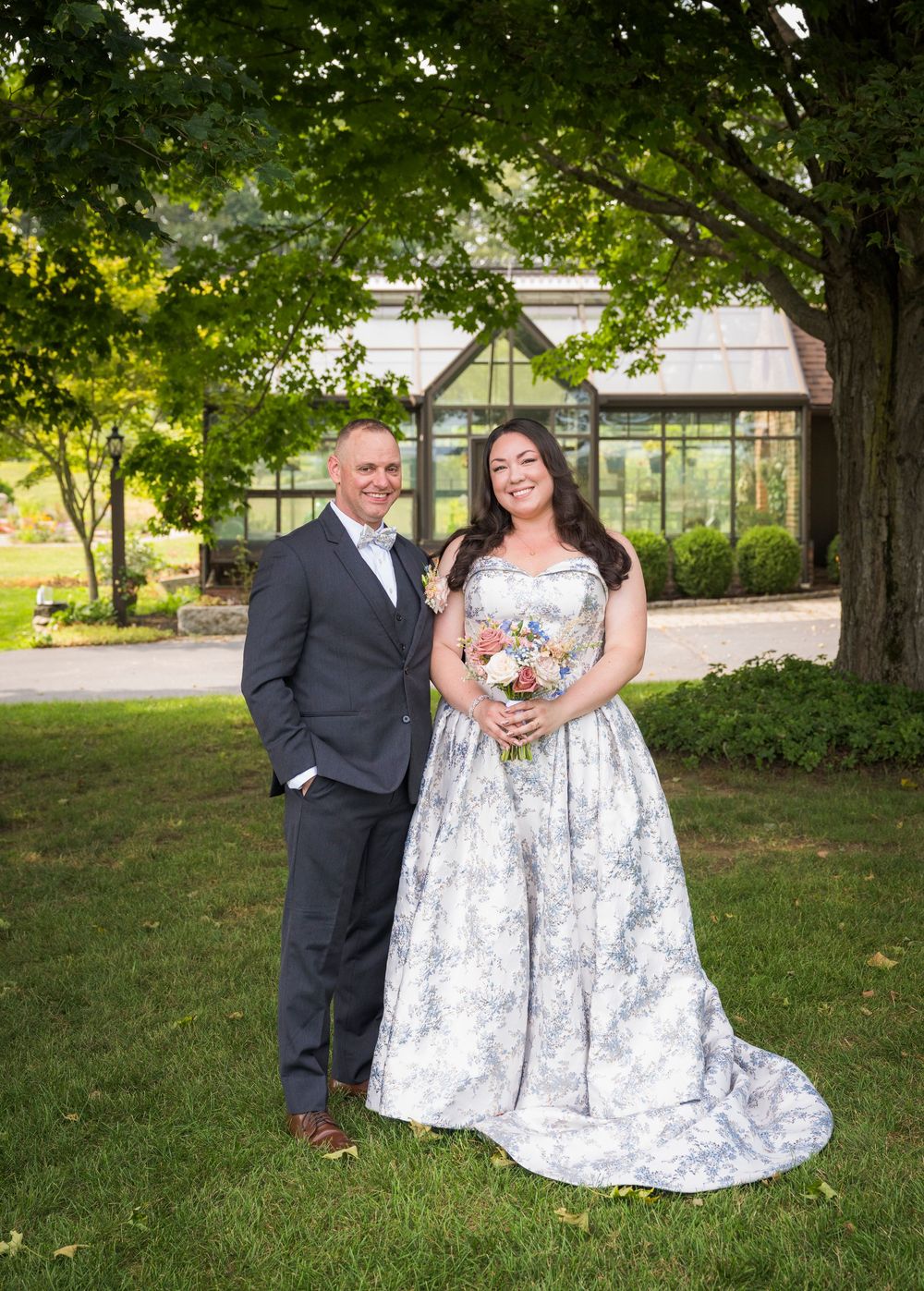 A wedding couple poses on a lush lawn with a glass greenhouse conservatory visible in the background on a sunny day.