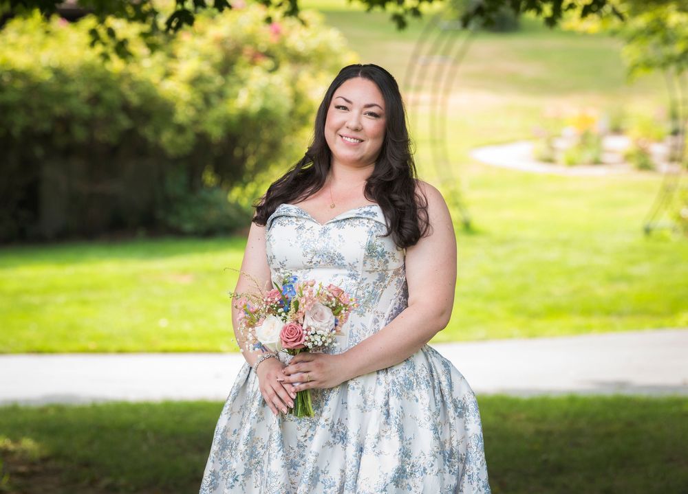 Row of smiling bridesmaids in floral blue dresses holding bouquets in sunny garden setting.
