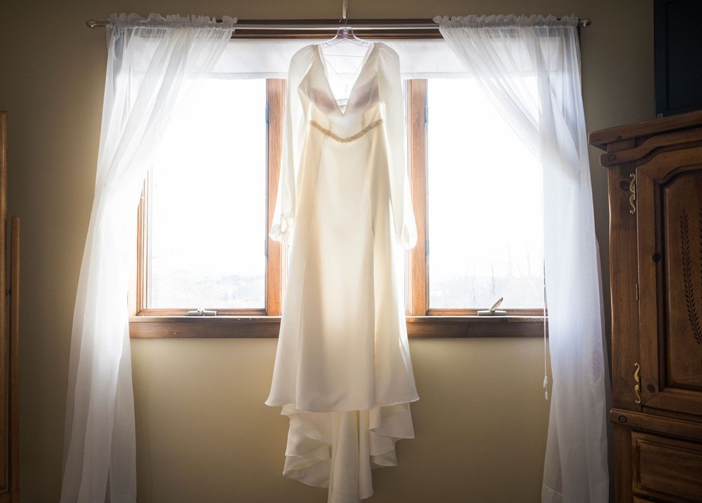 Wedding dress hanging in window with natural sunlight streaming through sheer curtains.