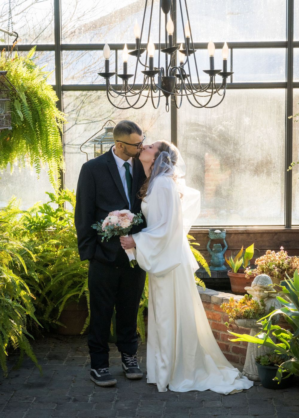 A couple shares an intimate moment in a sunlit greenhouse surrounded by lush ferns and hanging plants.