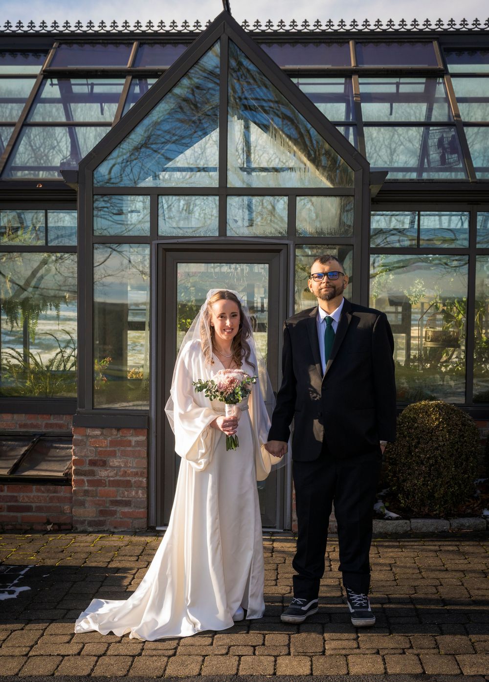 A couple poses in formal wedding attire in front of a glass conservatory on a brick patio.