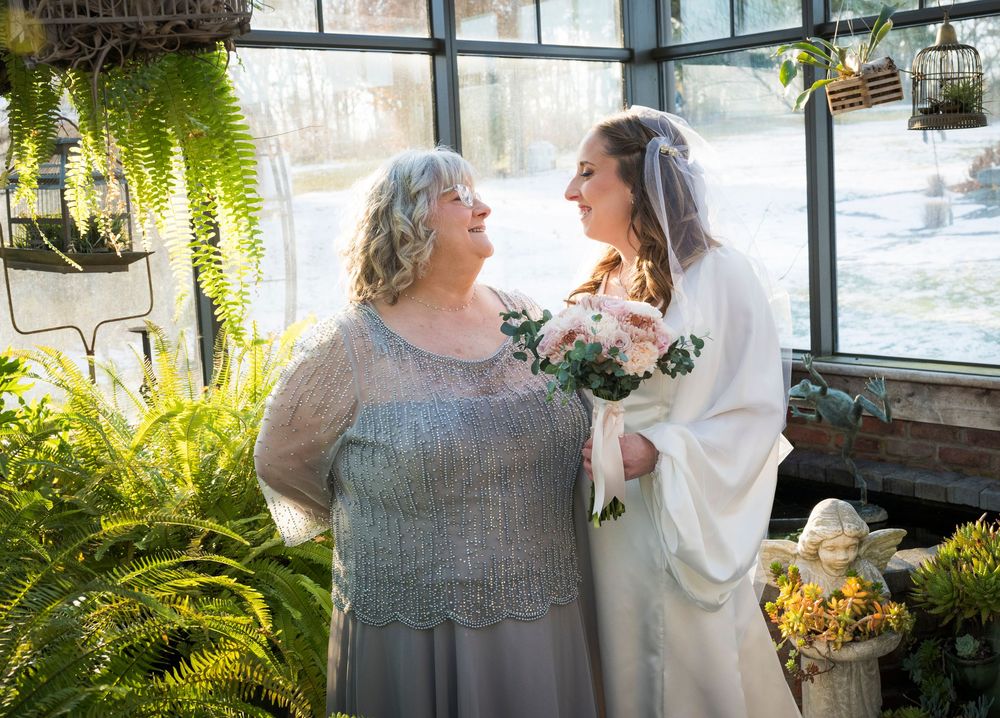 Two people share a joyful moment surrounded by ferns and greenery in a sunlit greenhouse setting.