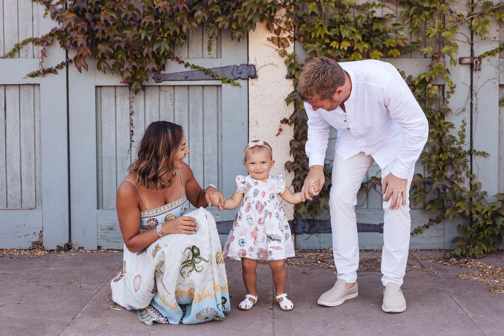 Family members wearing white outfits gather against a blue wall with climbing vines for a photo session.