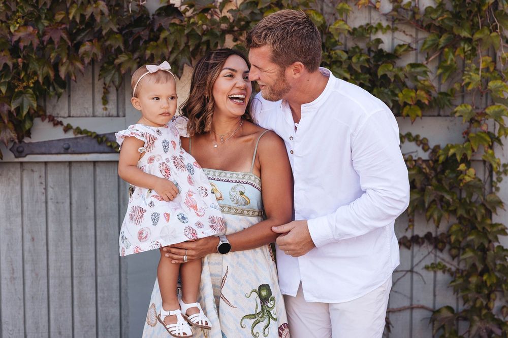 Family wearing coordinated white outfits share tender moments together against an ivy-covered wall background.