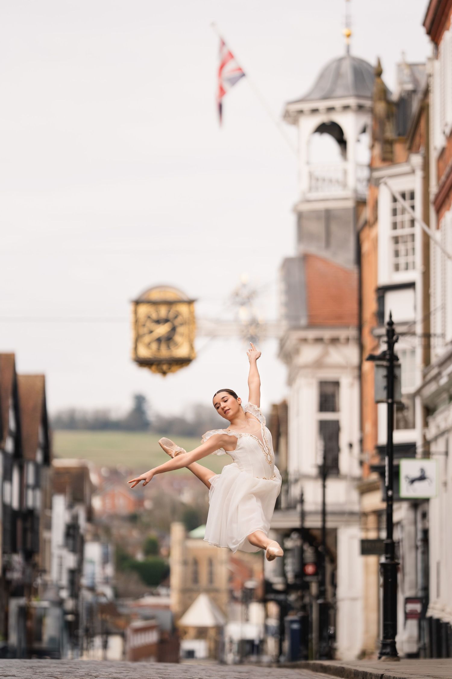 A ballet dancer performs a graceful leap on a historic English town street with Tudor buildings and a clock tower in view.