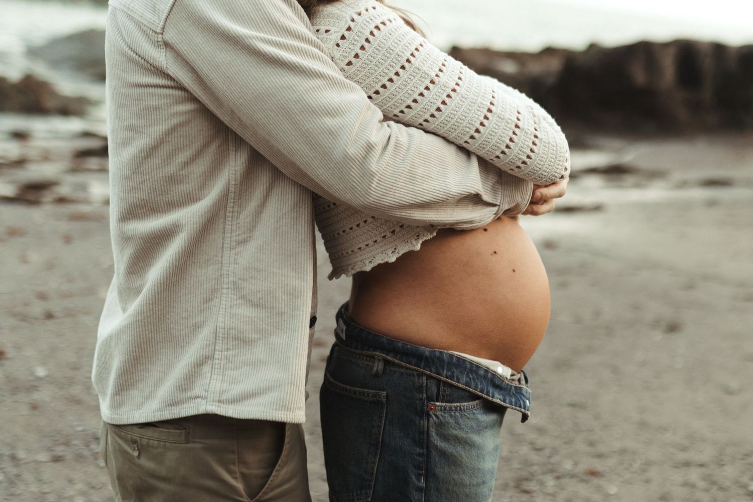 A romantic maternity photoshoot captured on a cloudy beach shows an expectant couple in casual attire embracing.
