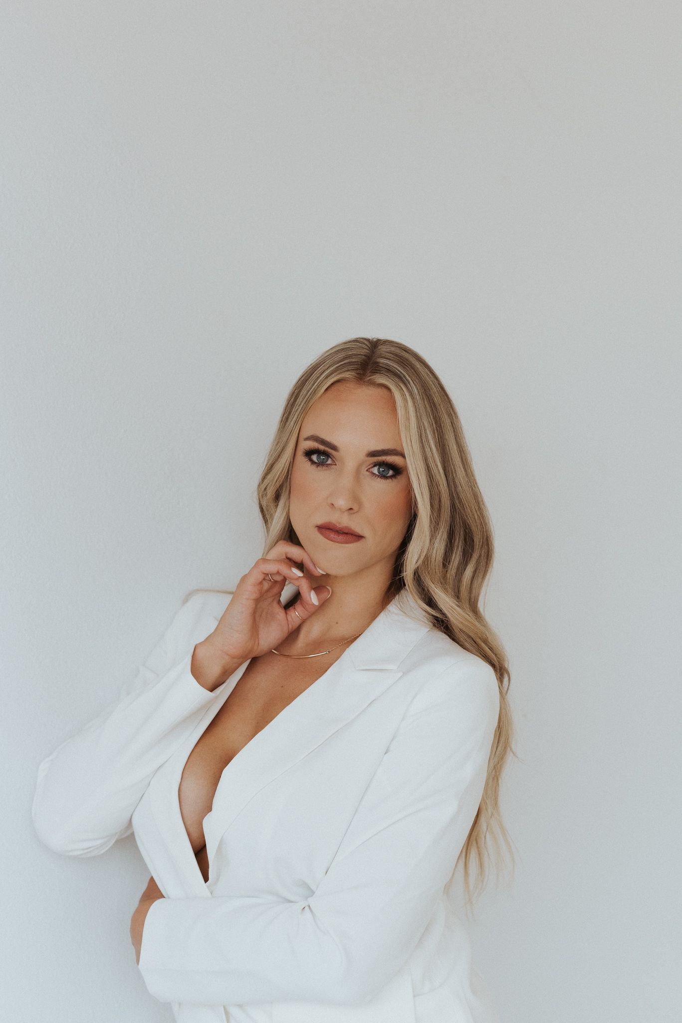 Woman in white blazer poses elegantly against plain white wall.