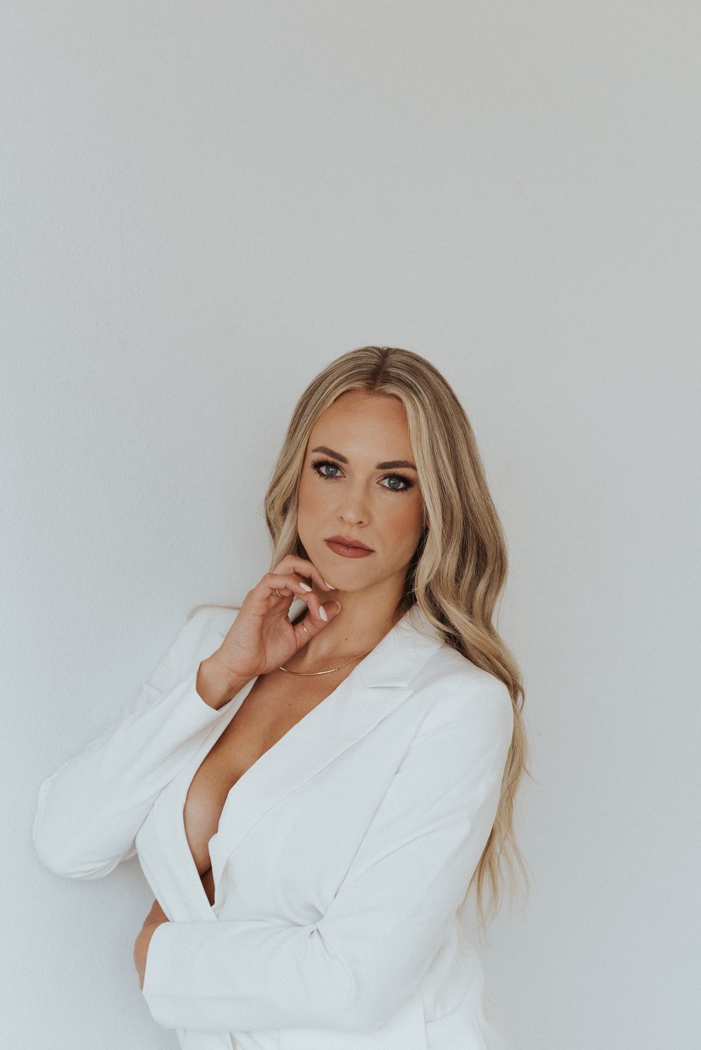 Woman in white blazer poses elegantly against plain white wall.