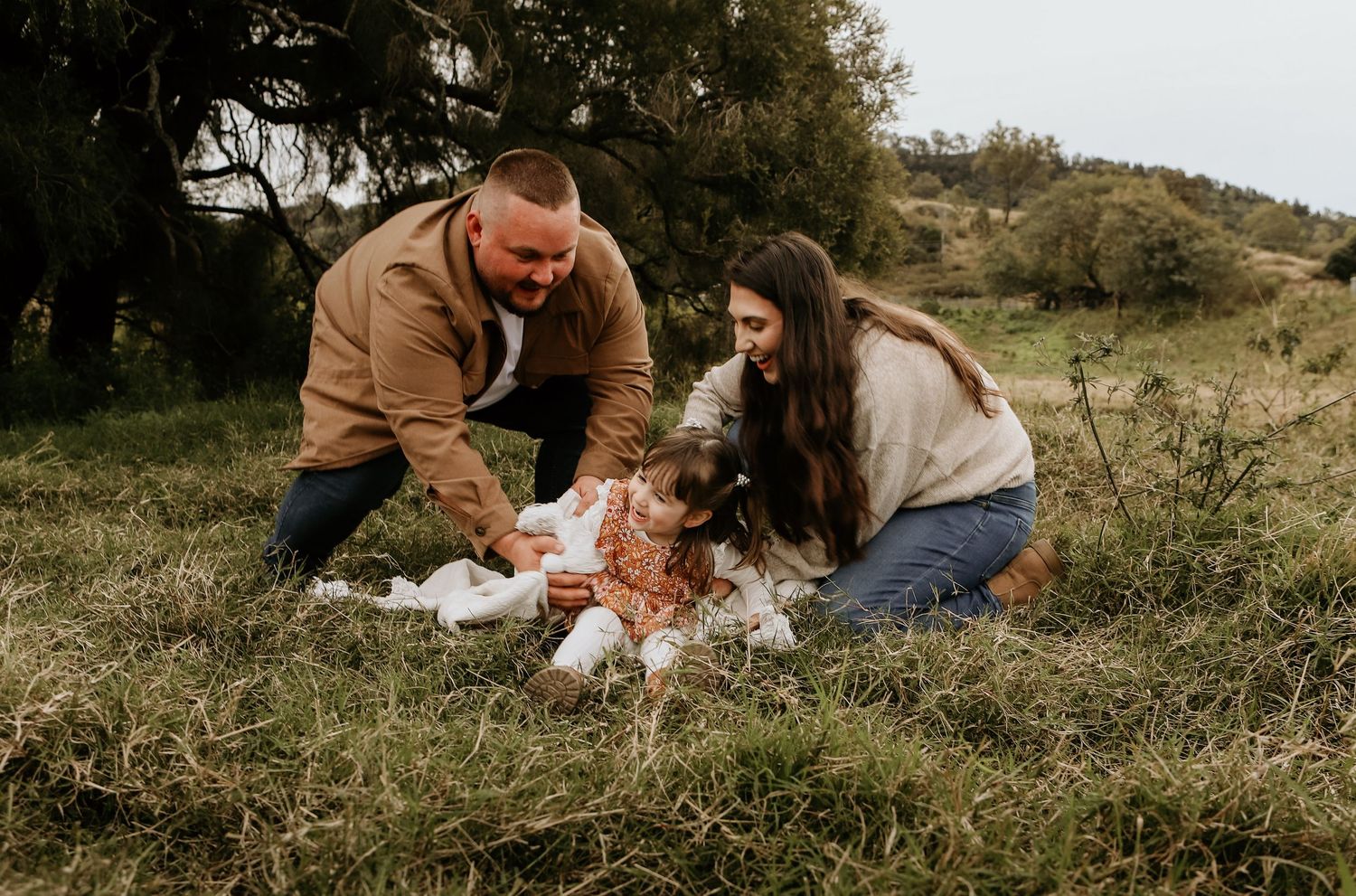 A couple spends time with their baby on a blanket in a grassy hillside on an overcast day.