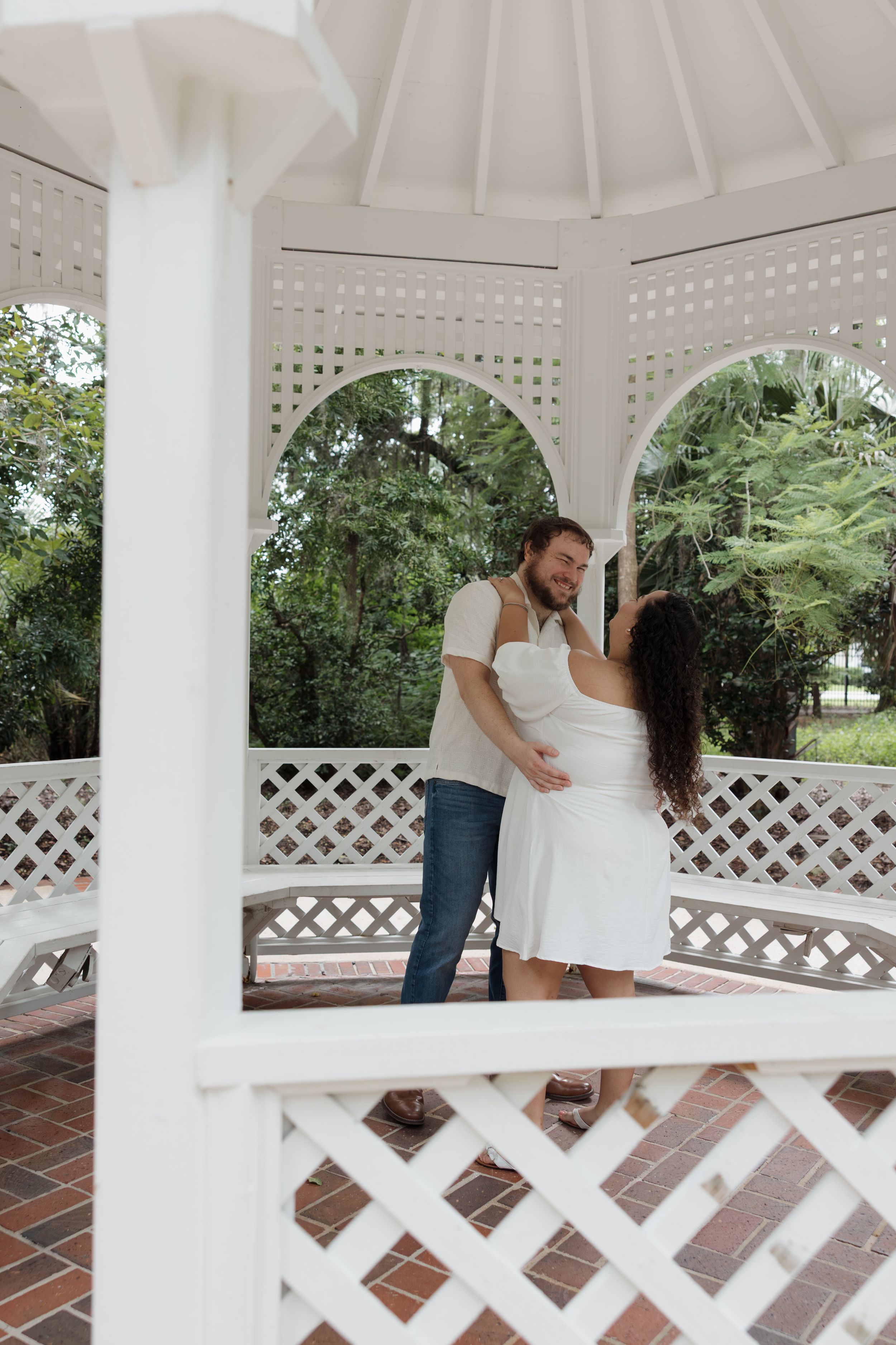 A couple shares an intimate moment inside a white Victorian-style gazebo with lattice details.