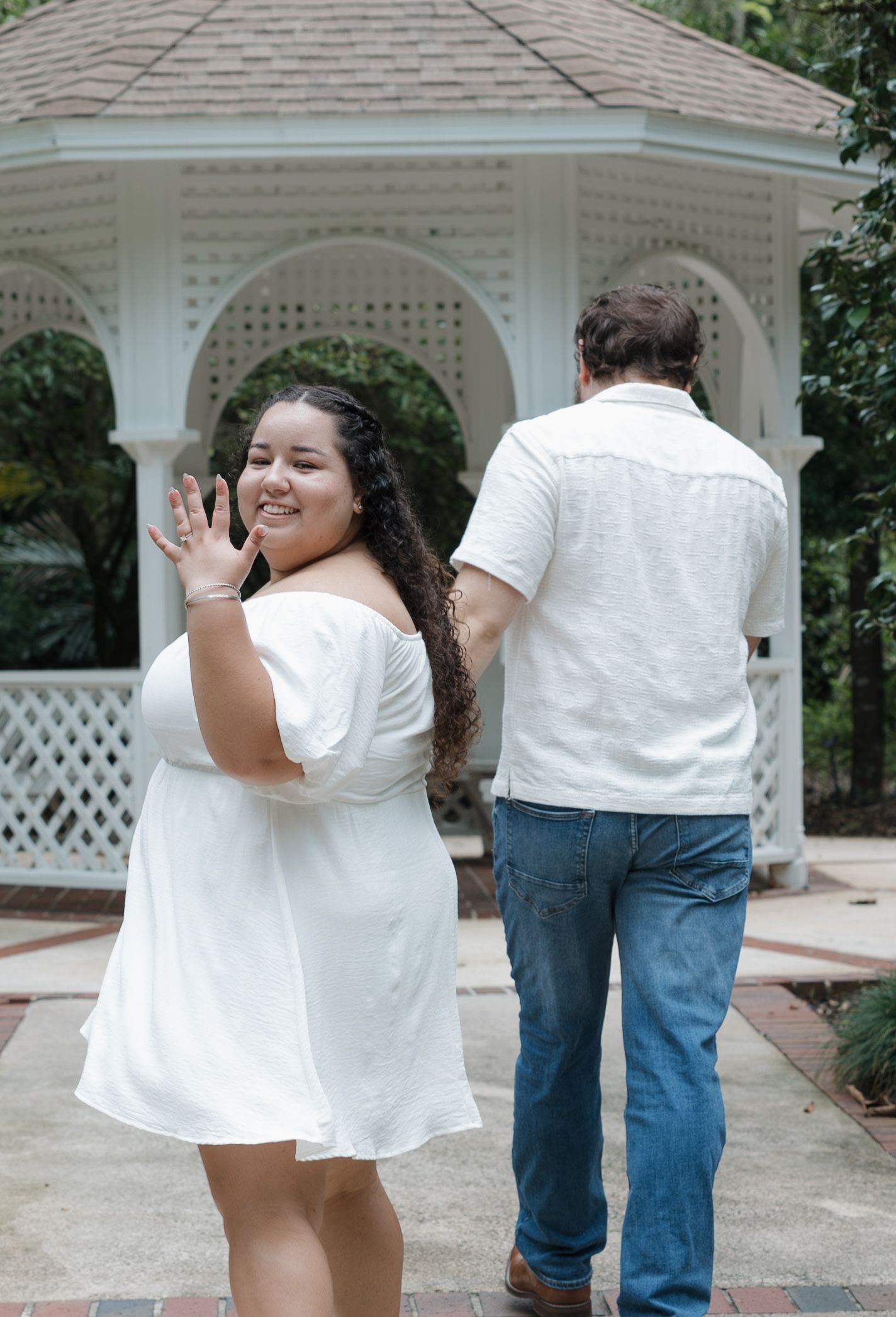 A couple embraces in front of a white gazebo in an outdoor garden setting.