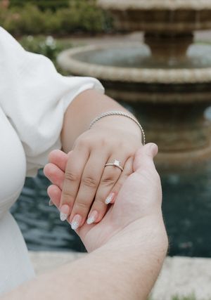 Couple holding hands showing engagement rings near a fountain.