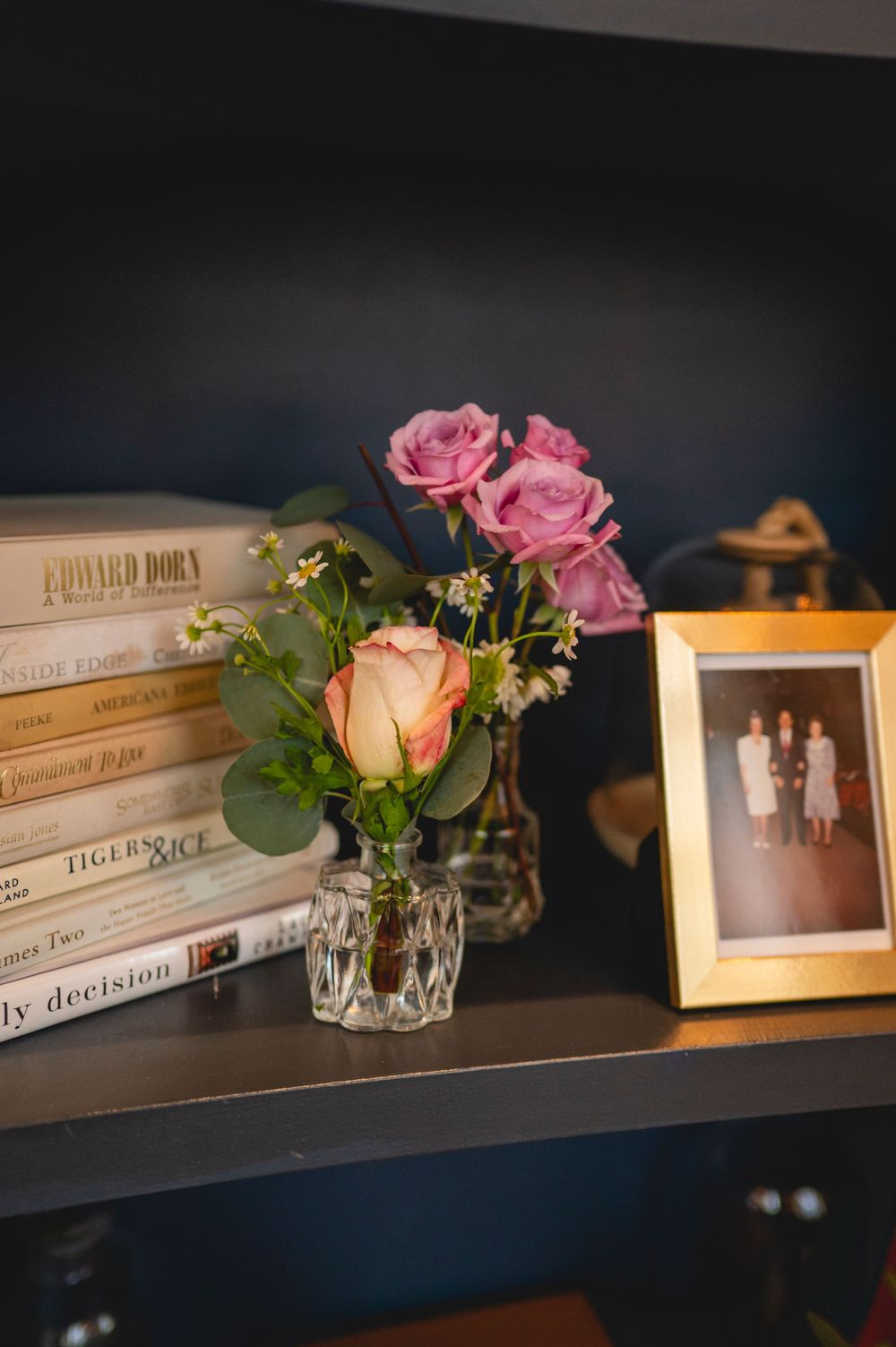 Books and pink roses in small vases line a dark wooden shelf with decorative photo frames.