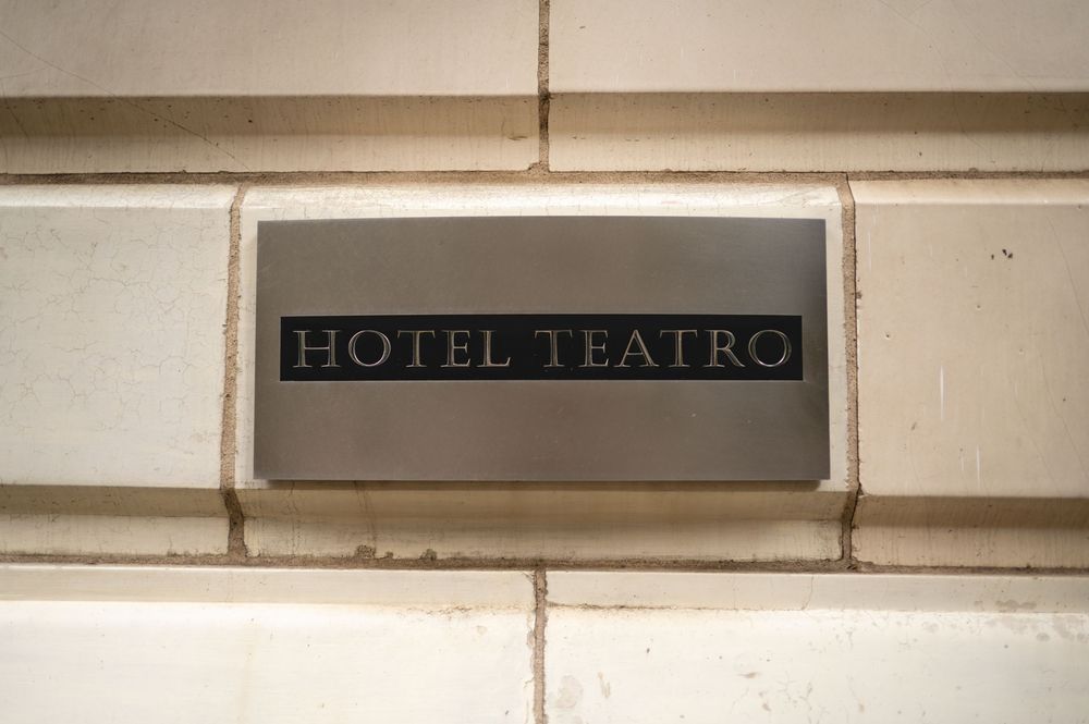 Black and white plaque reading 'HOTEL TEATRO' mounted on light colored stone wall with horizontal grout lines.