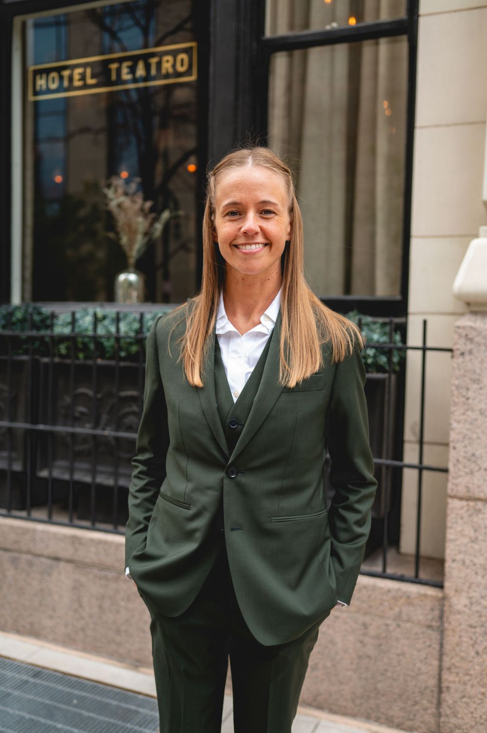 Professional wearing a dark green suit and white shirt stands outside Hotel Teatro with a welcoming expression.