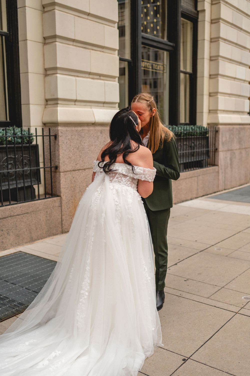 A bride in a flowy off-shoulder white gown shares a romantic moment with partner in dark suit on urban sidewalk.