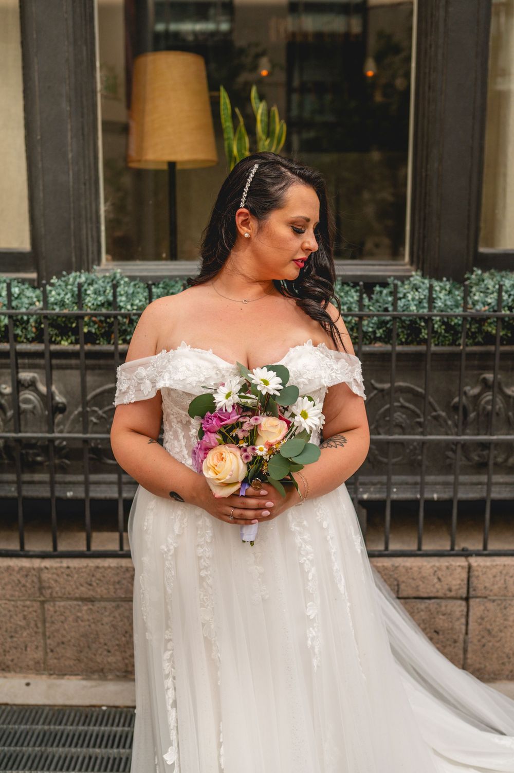 A bride wearing an off-shoulder white wedding gown holding a colorful bouquet stands in front of Hotel Teatro.