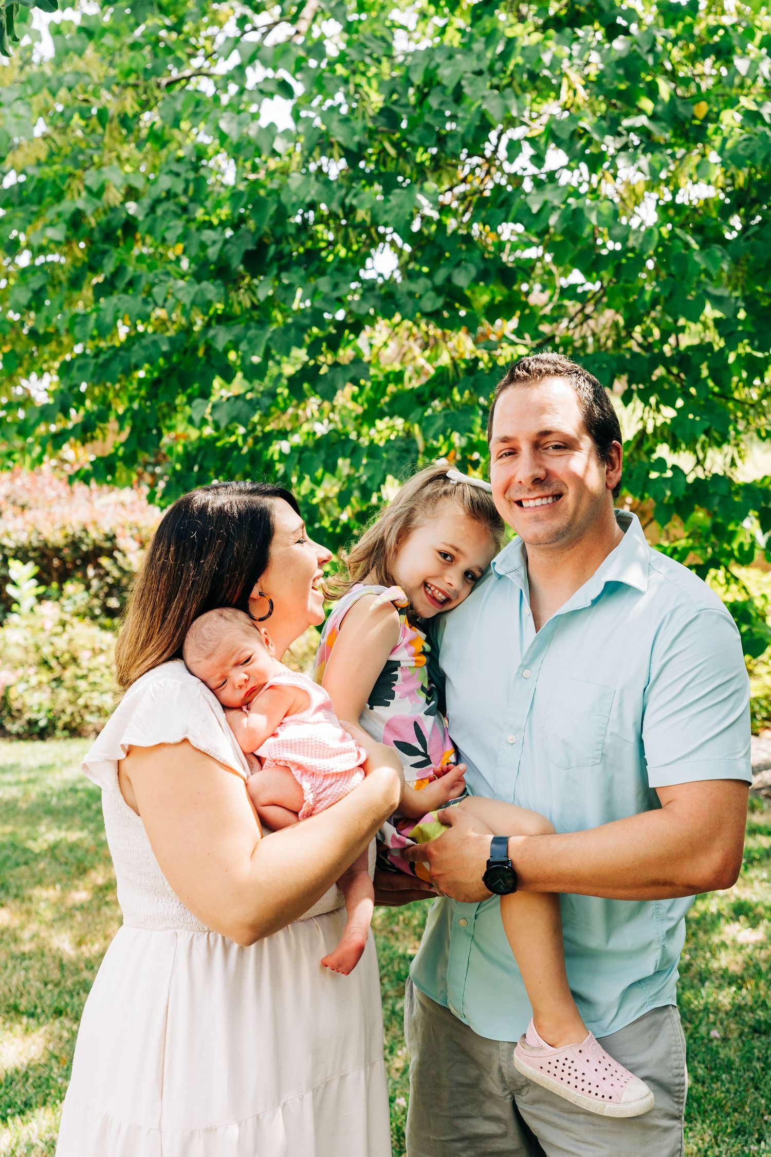 A family shares joyful laughter together outdoors on a sunny summer day with leafy green trees in the background.