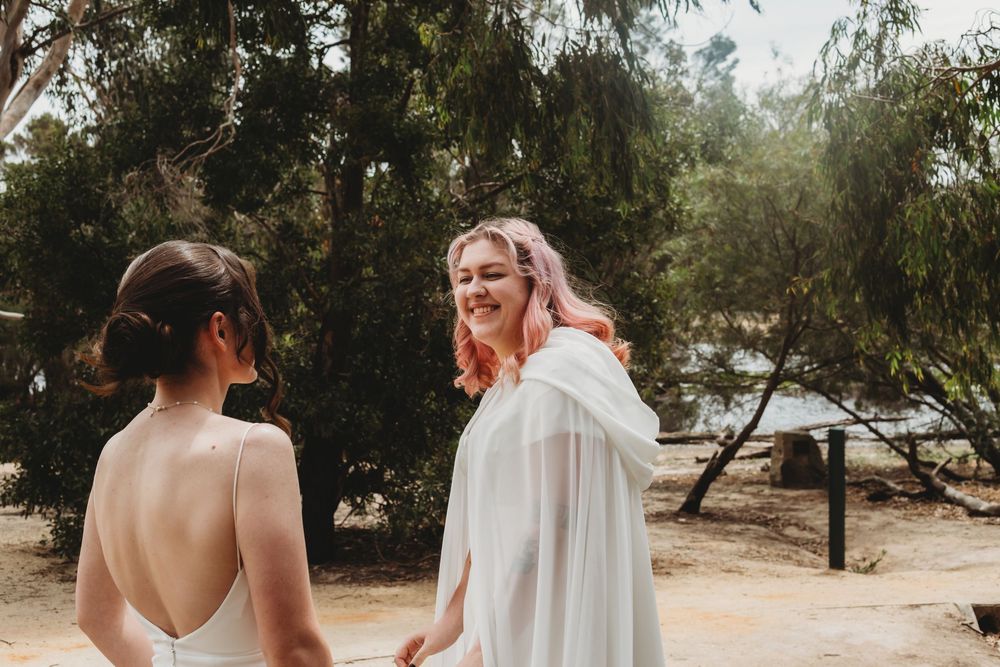 Two people share a joyful moment in a white dress and cape on a sunny day in an outdoor garden setting.