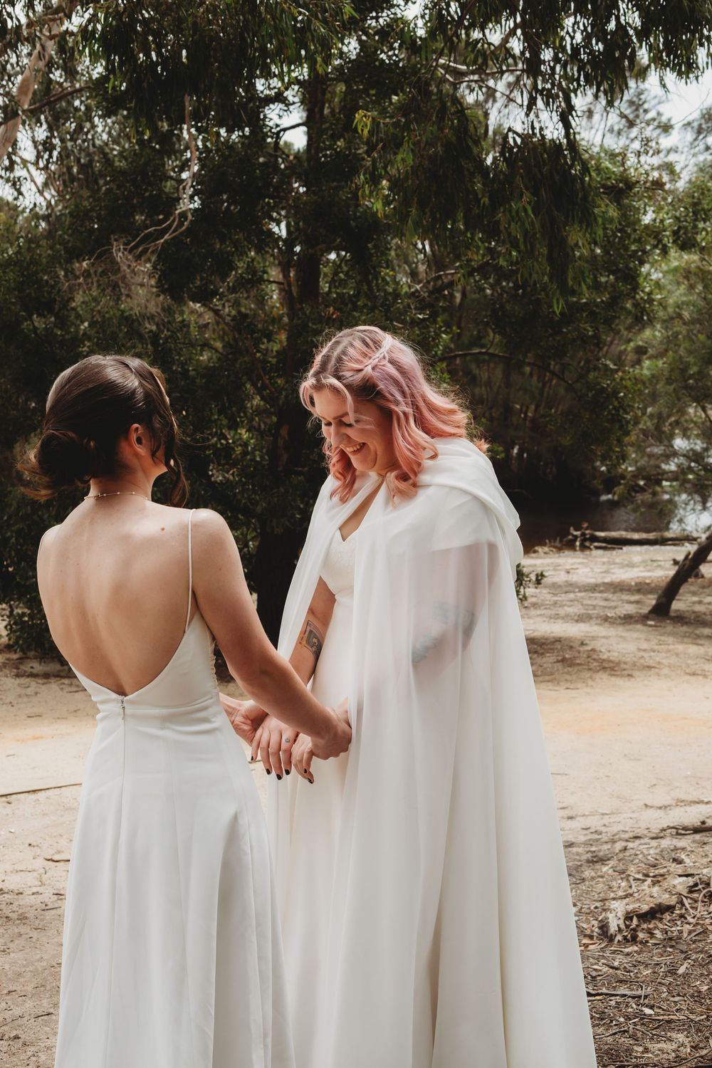 Two people wearing white wedding dresses walking together on a wooded path outdoors in soft natural lighting.