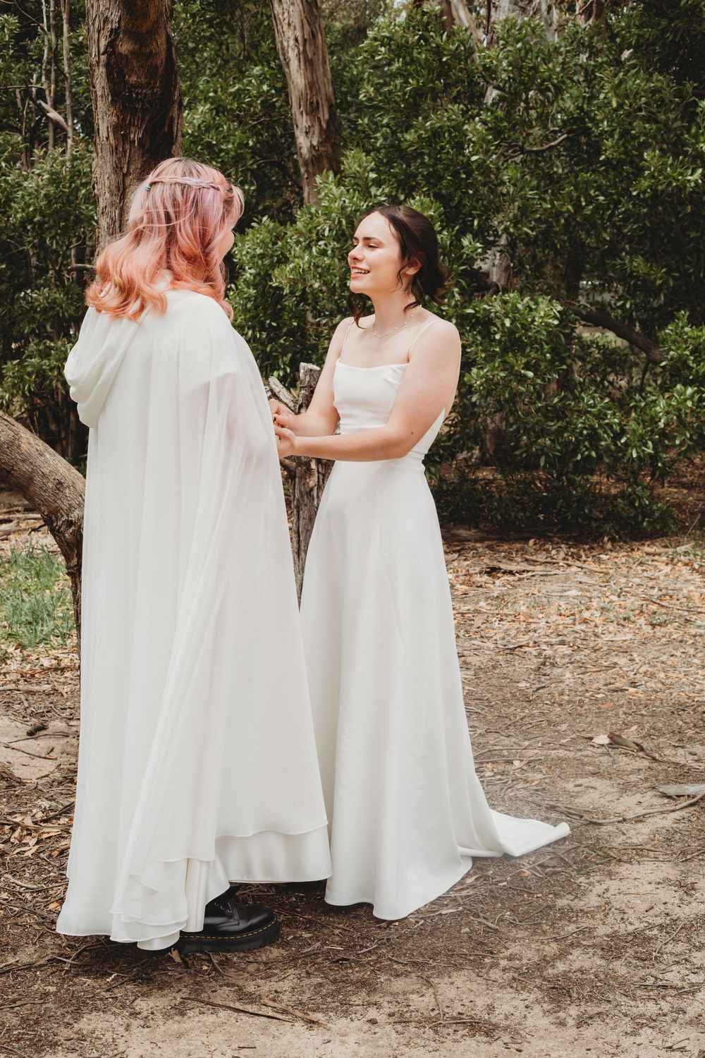 Two people in flowing white wedding gowns share a candid laughing moment together in an outdoor wooded setting.