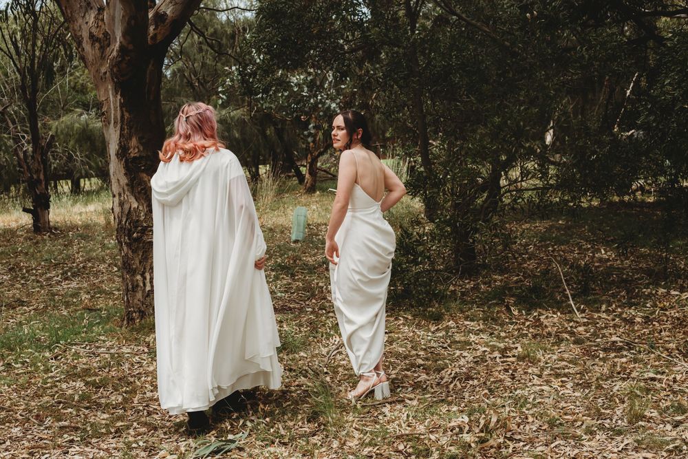 Two figures in white dresses walk through fallen leaves in a wooded outdoor setting.