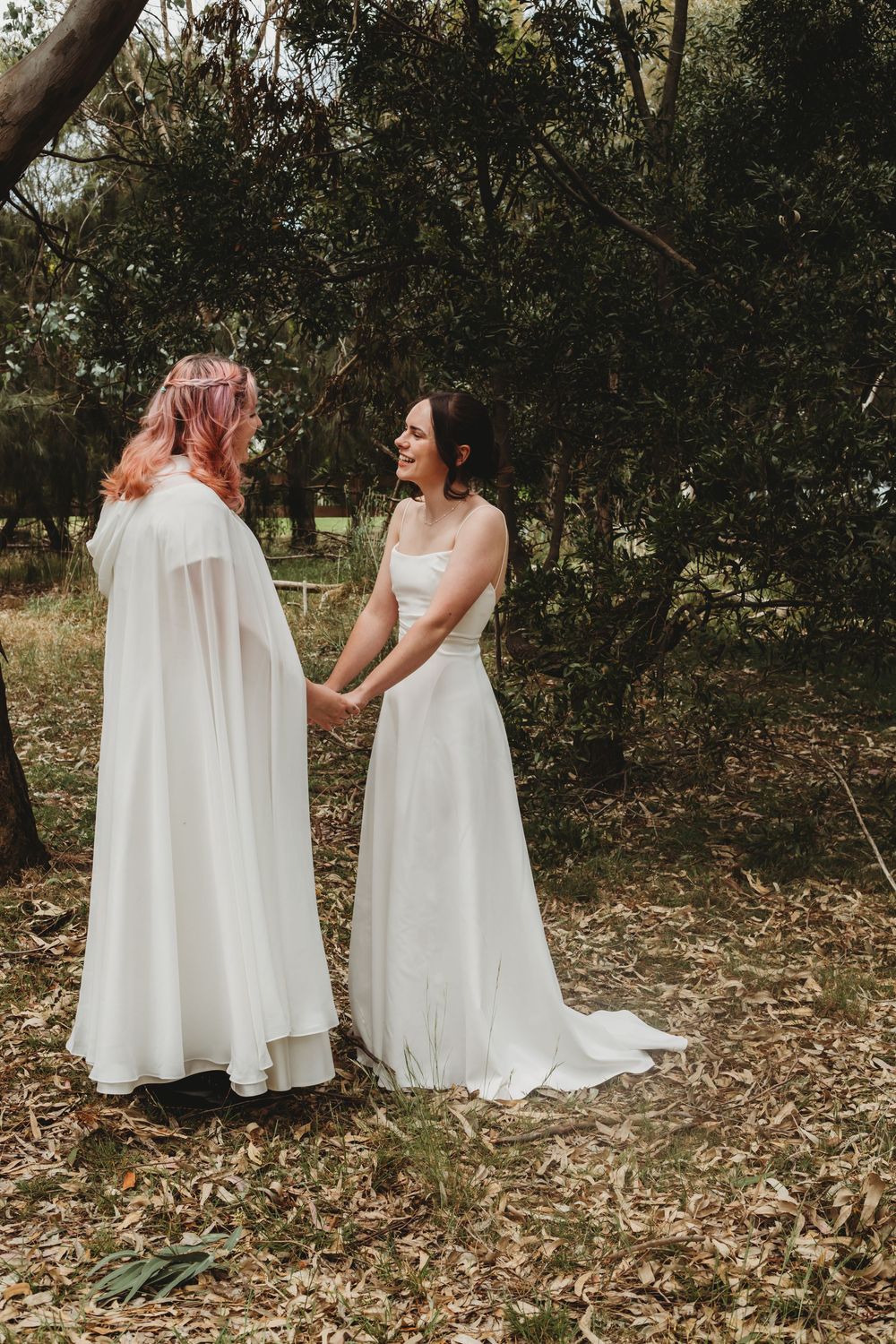 Two people in white wedding dresses stand together in a wooded outdoor setting during a romantic photo session.