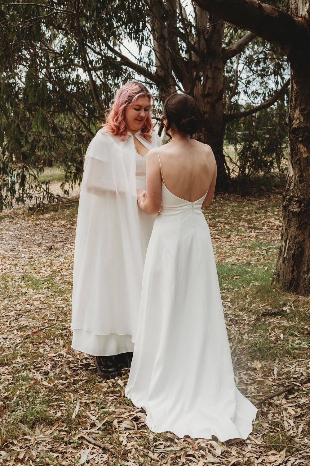 Two people in elegant white dresses walk among eucalyptus trees in an outdoor woodland setting.