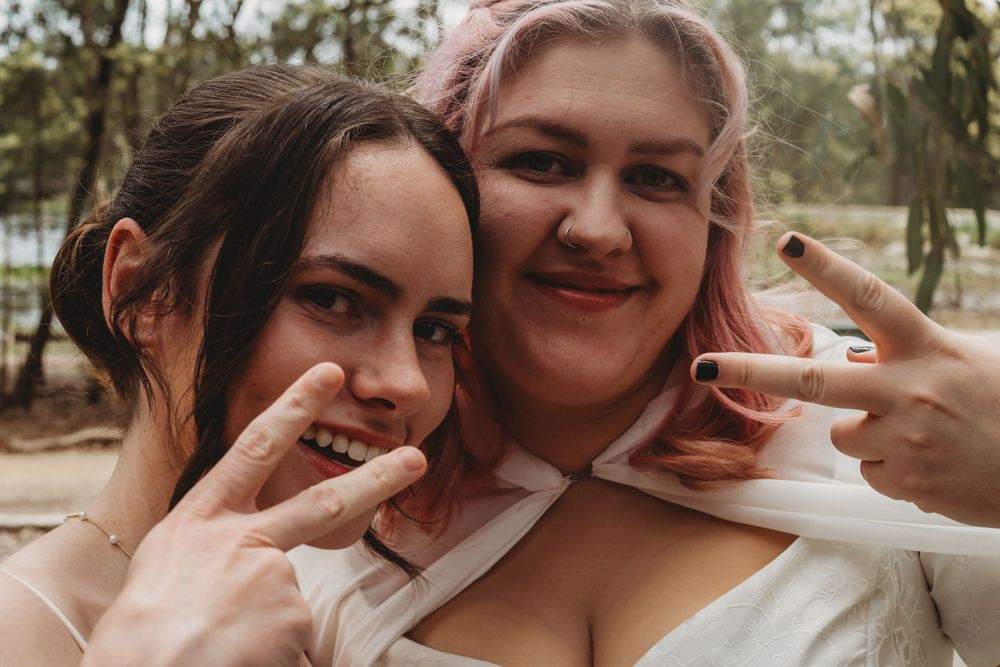 Close-up candid portrait of two smiling friends with pink hair making peace signs.