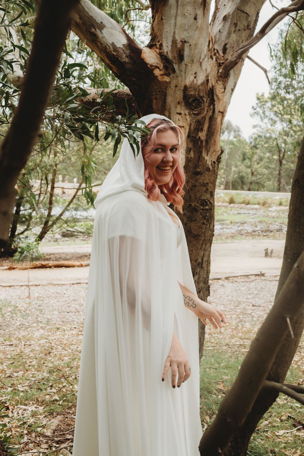 Beautiful bride in flowing white dress and cape stands gracefully among eucalyptus trees in an outdoor setting.