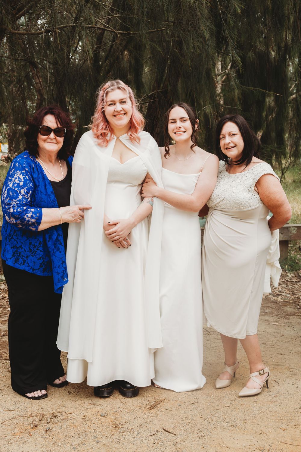 Four women pose together for a wedding photo outdoors wearing white and cream dresses with one in a blue lace shawl.