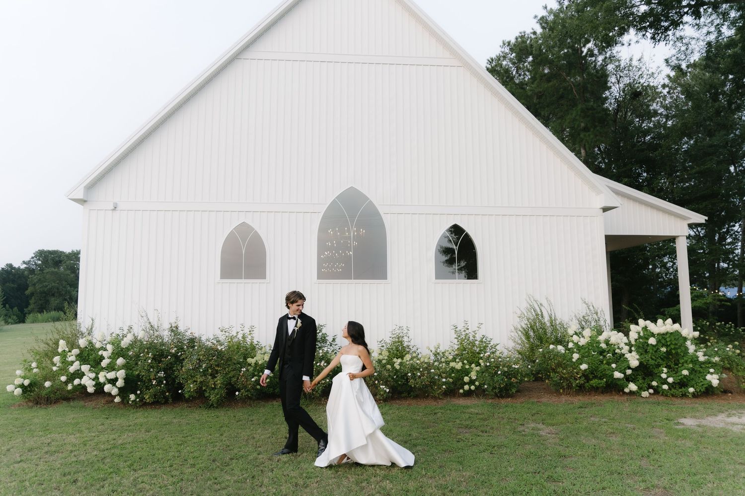 Bride and groom walking hand in hand in front of a white chapel surrounded by lush greenery on their wedding day.