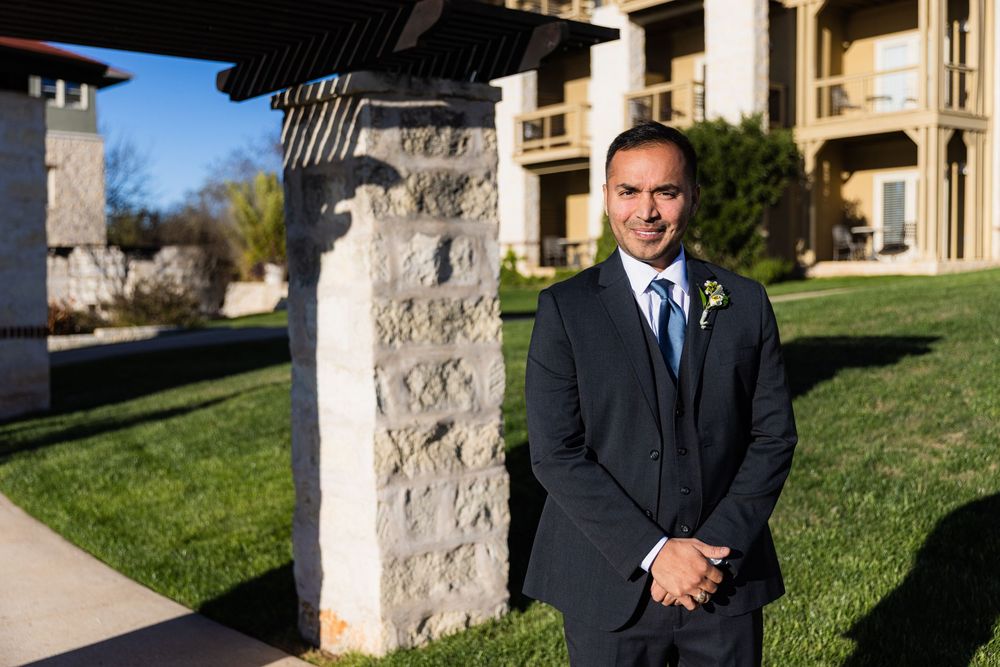 A person in a dark business suit poses near stone pillars with arched architecture and green lawn in the background.