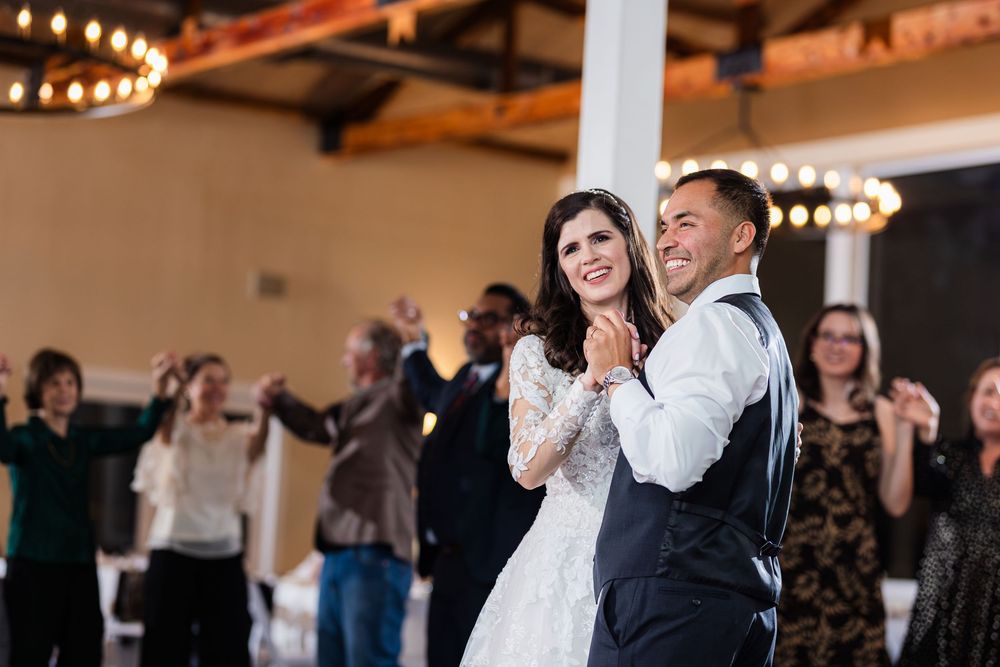 Wedding guests in a dimly lit reception venue dance in a group under hanging string lights.