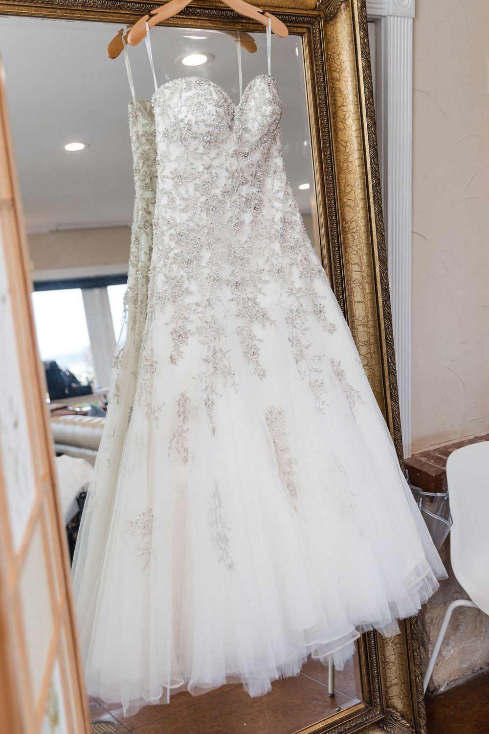 Elegant white wedding dress with lace details and full tulle skirt displayed in an ornate gold framed mirror.