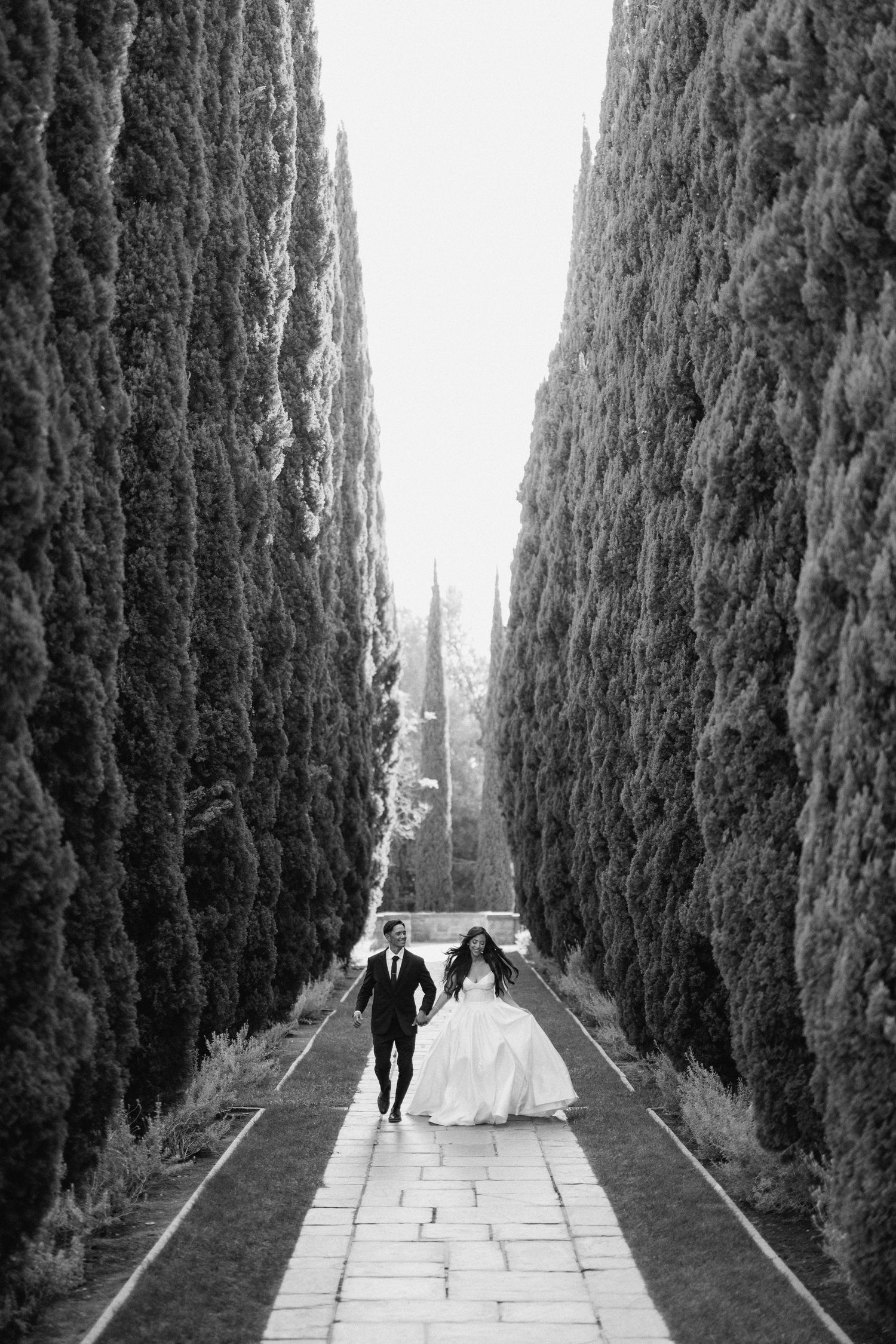 People walk down a cypress tree-lined path in a formal garden captured in four sequential black and white frames.