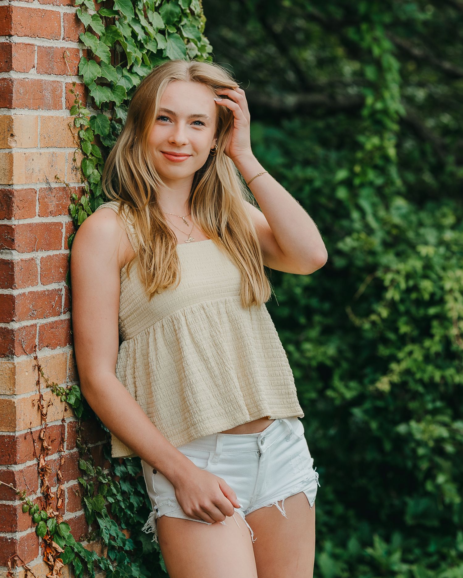 A senior girl in a beige top and white shorts poses casually against a brick wall with green foliage in the background.