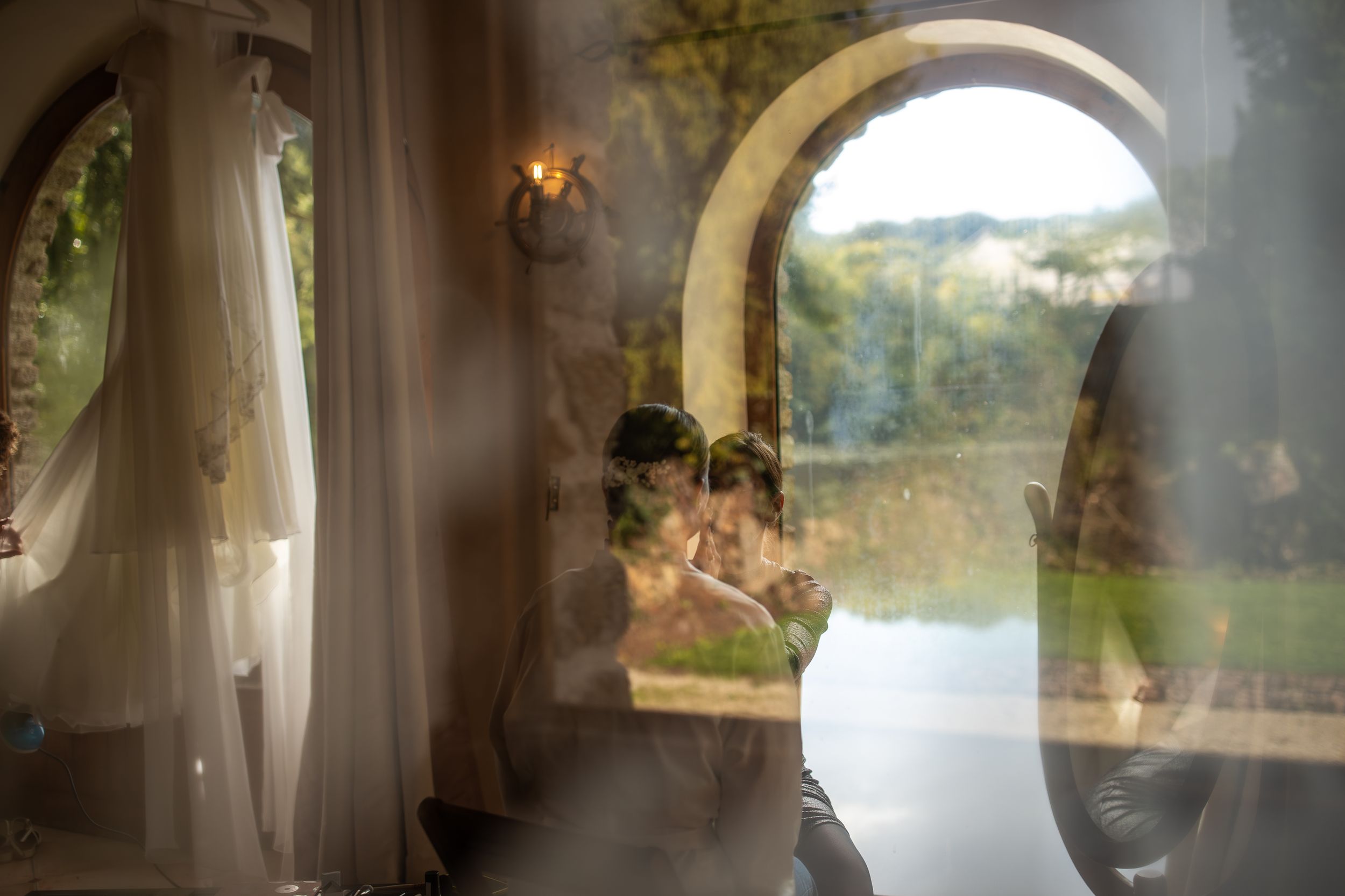 reflet d'une femme qui se fait maquiller par son amie le jour de son mariage près d'un lac à travers une fenêtre