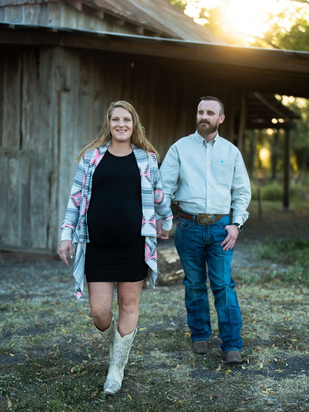 A couple stands together by a weathered barn at sunset wearing western-inspired outfits.