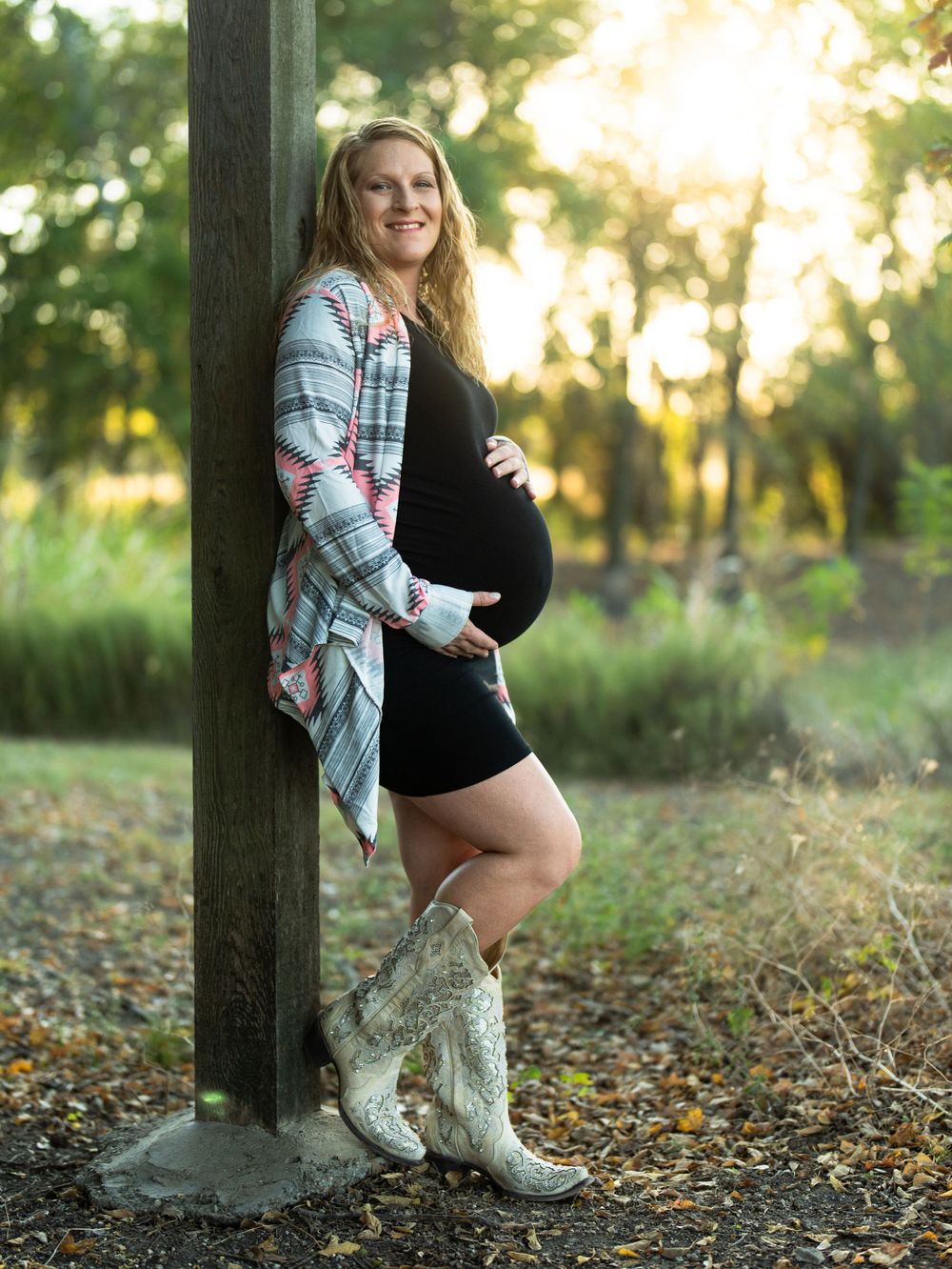Maternity portrait featuring cowboy boots and patterned cardigan against wooden post at sunset.