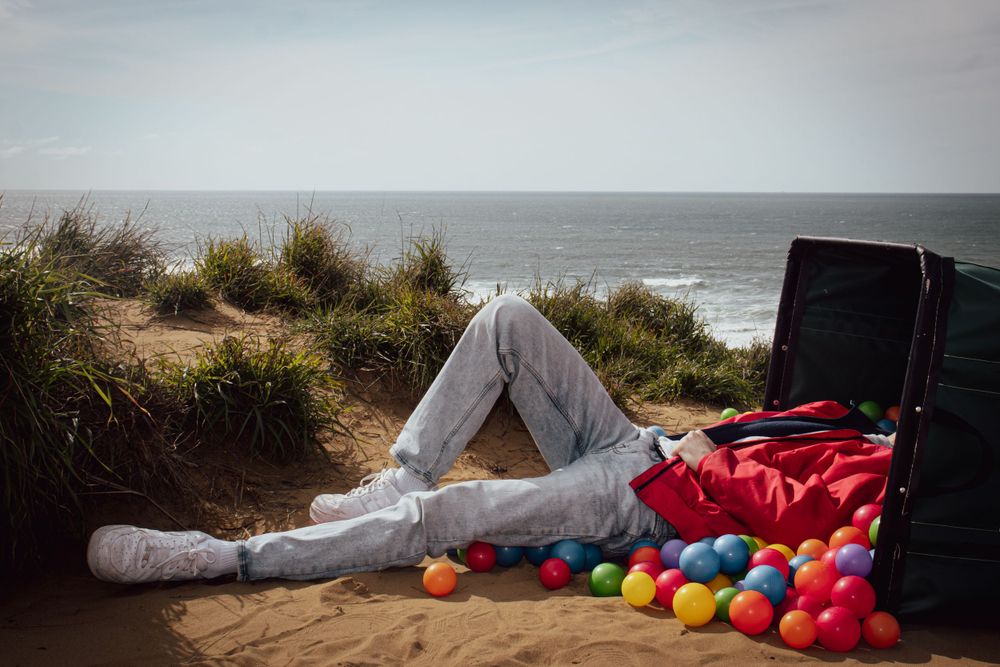 Colorful plastic balls scattered on beach near black case with person resting nearby.