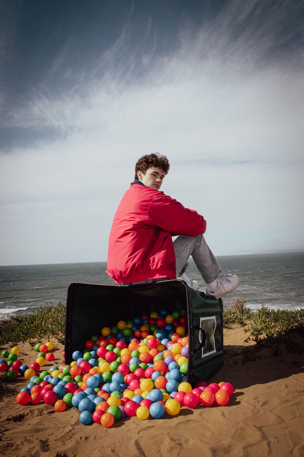 Person in red jacket sits on case surrounded by colorful balls on coastal overlook.