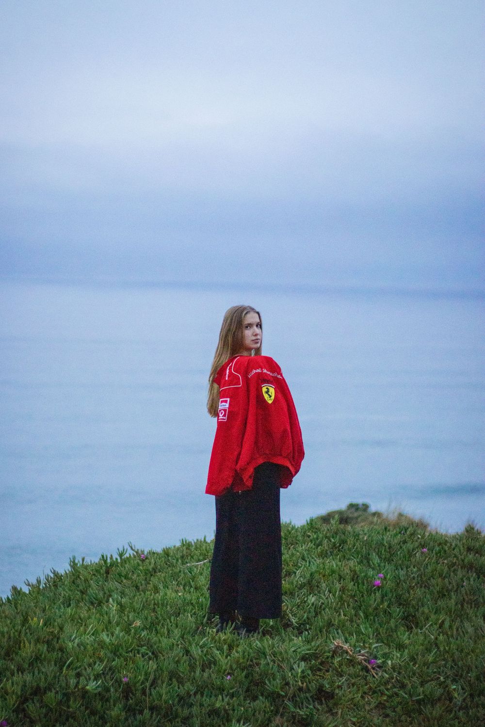 Person in red jacket stands on grassy hilltop overlooking ocean under cloudy sky at dusk.