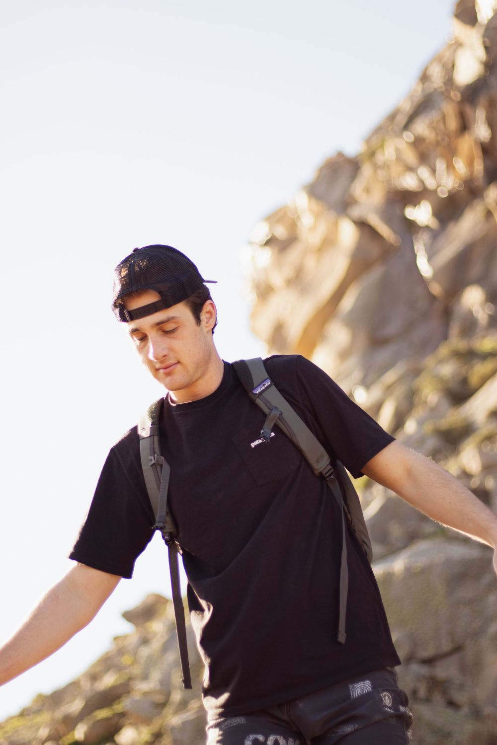 Hiker wearing black t-shirt and backpack explores rocky terrain at sunset.