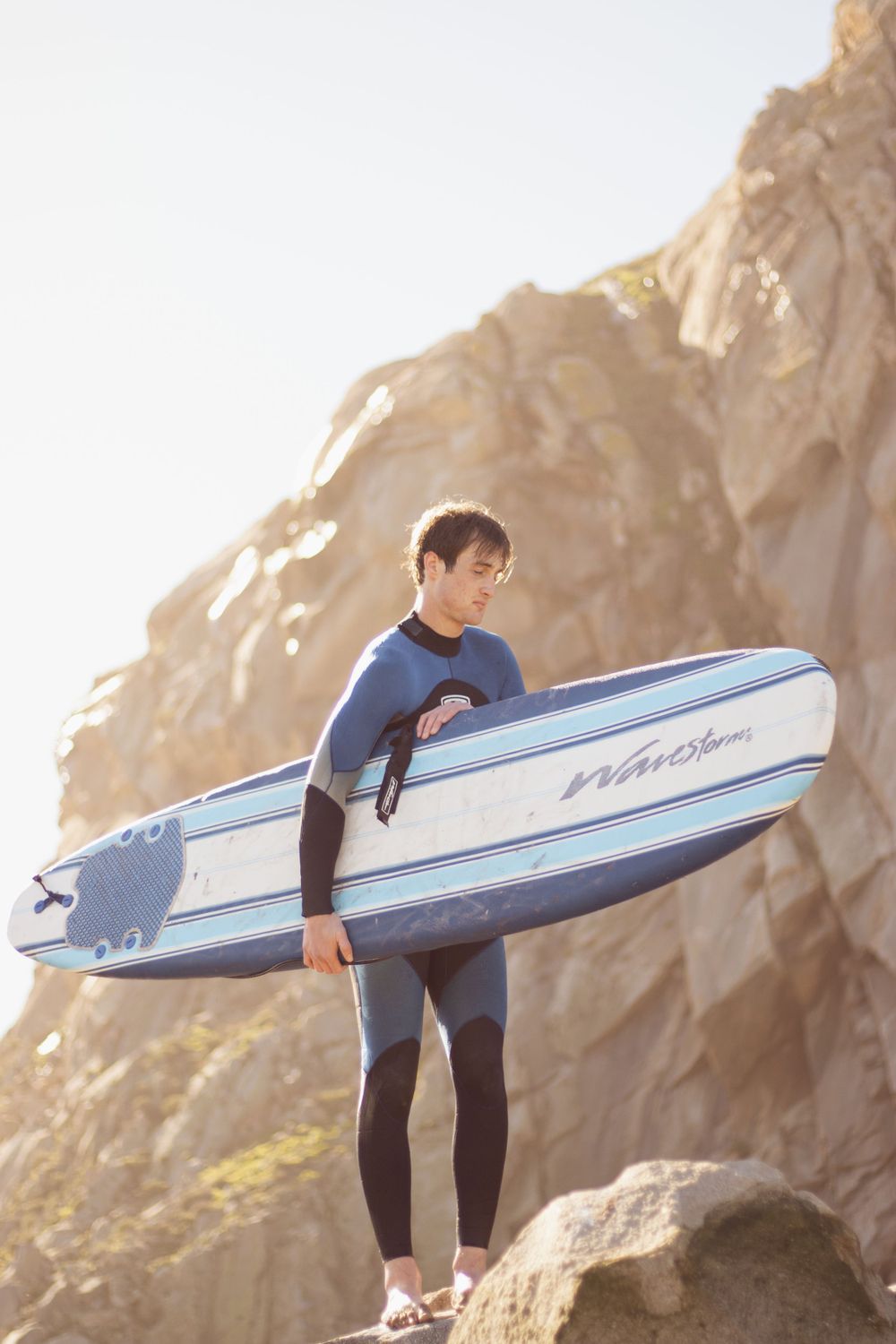 Surfer carrying blue and white surfboard stands on rocky coastal cliff.