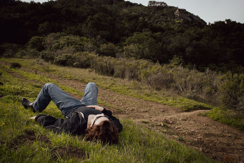 Person lying down peacefully on a grassy hill trail surrounded by nature.