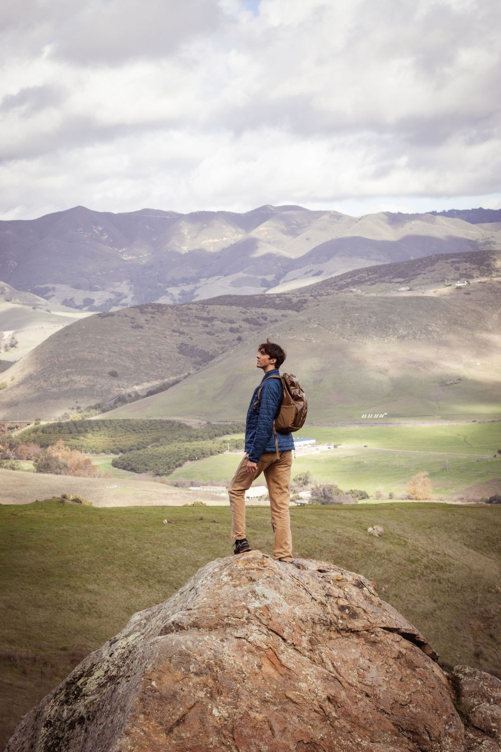 Adventurer stands on rocky outcrop overlooking vast mountain valley landscape.