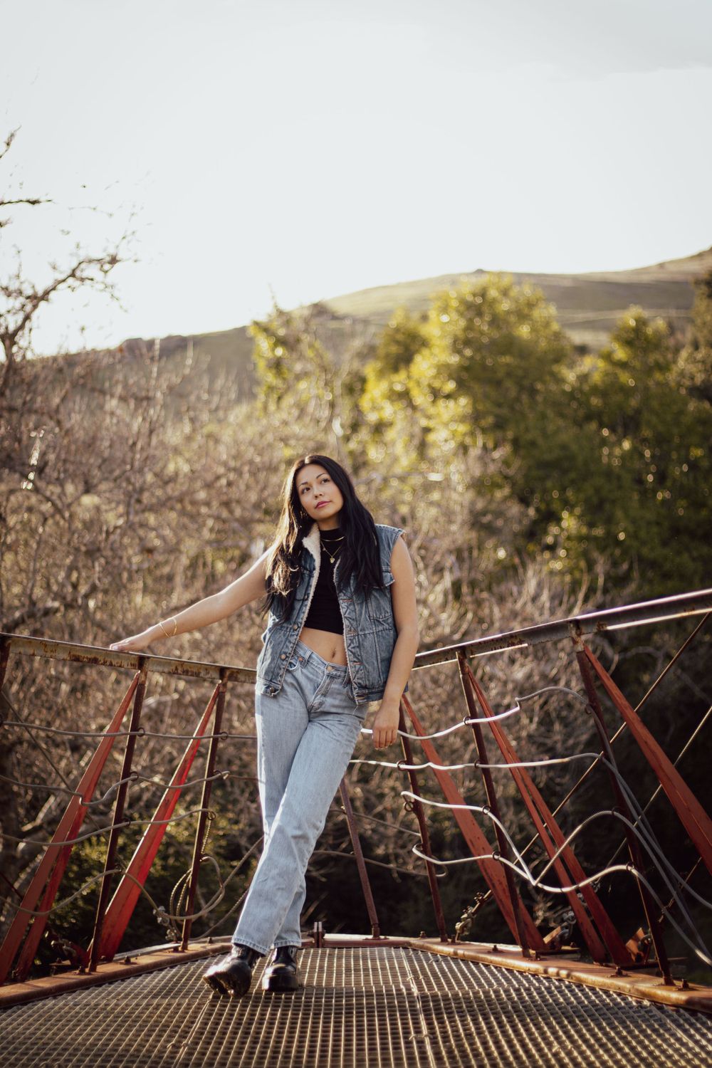 Figure in denim outfit posing on metal bridge structure with hills in background.