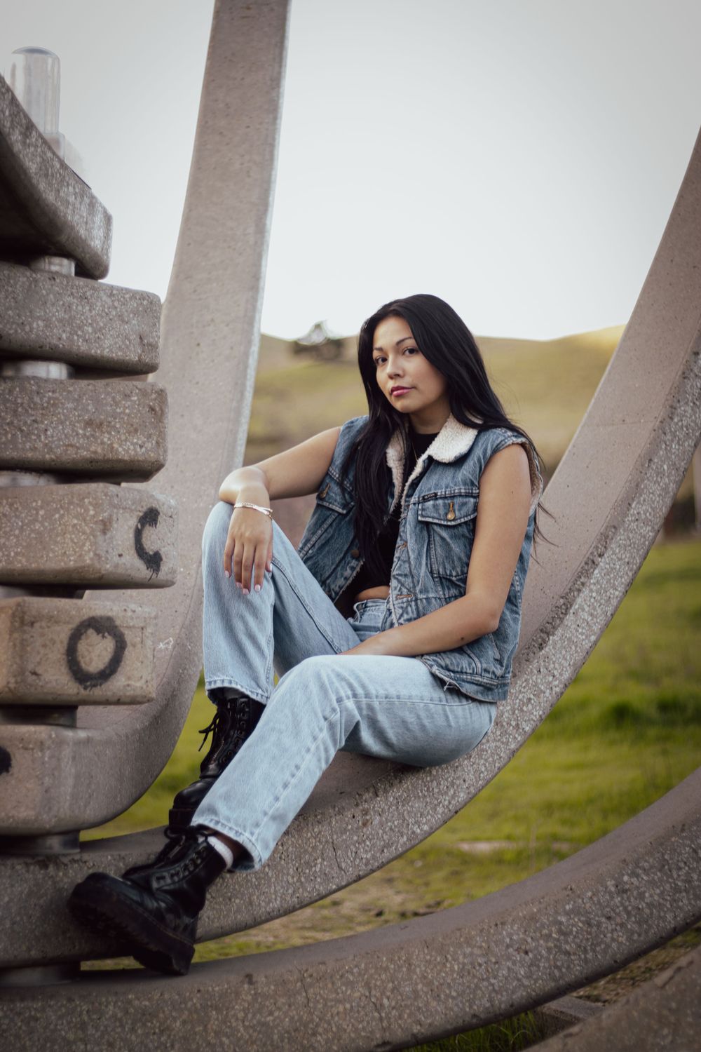 Person in denim vest and jeans sitting on concrete structure outdoors.