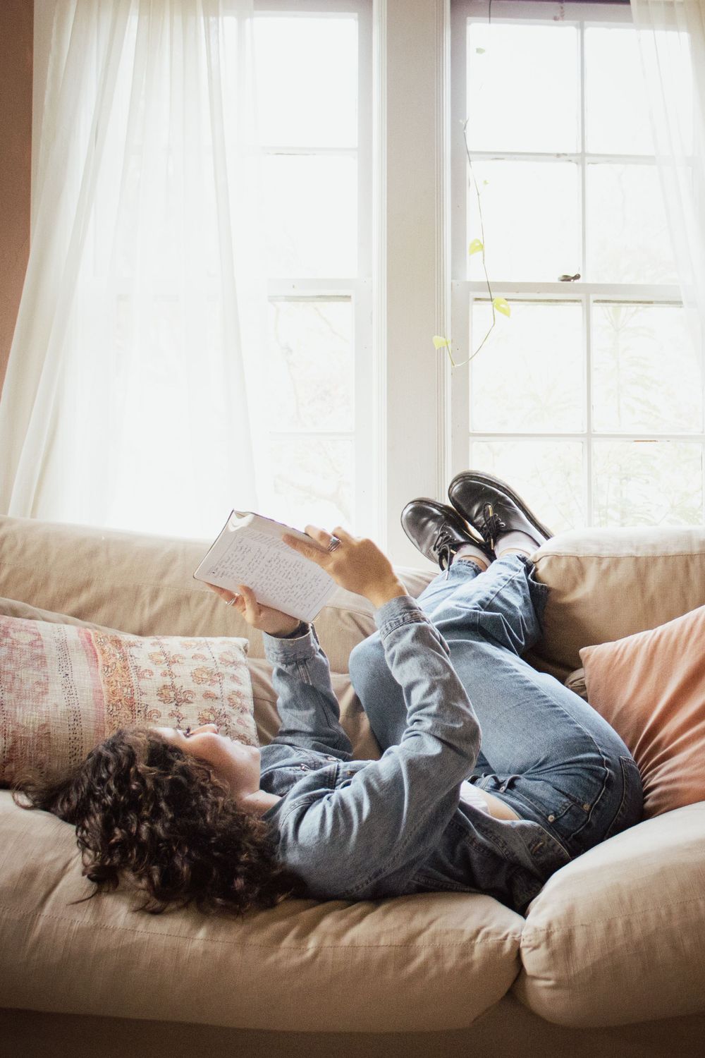 Person relaxing on a couch with a book by a sunny window in a casual indoor setting.